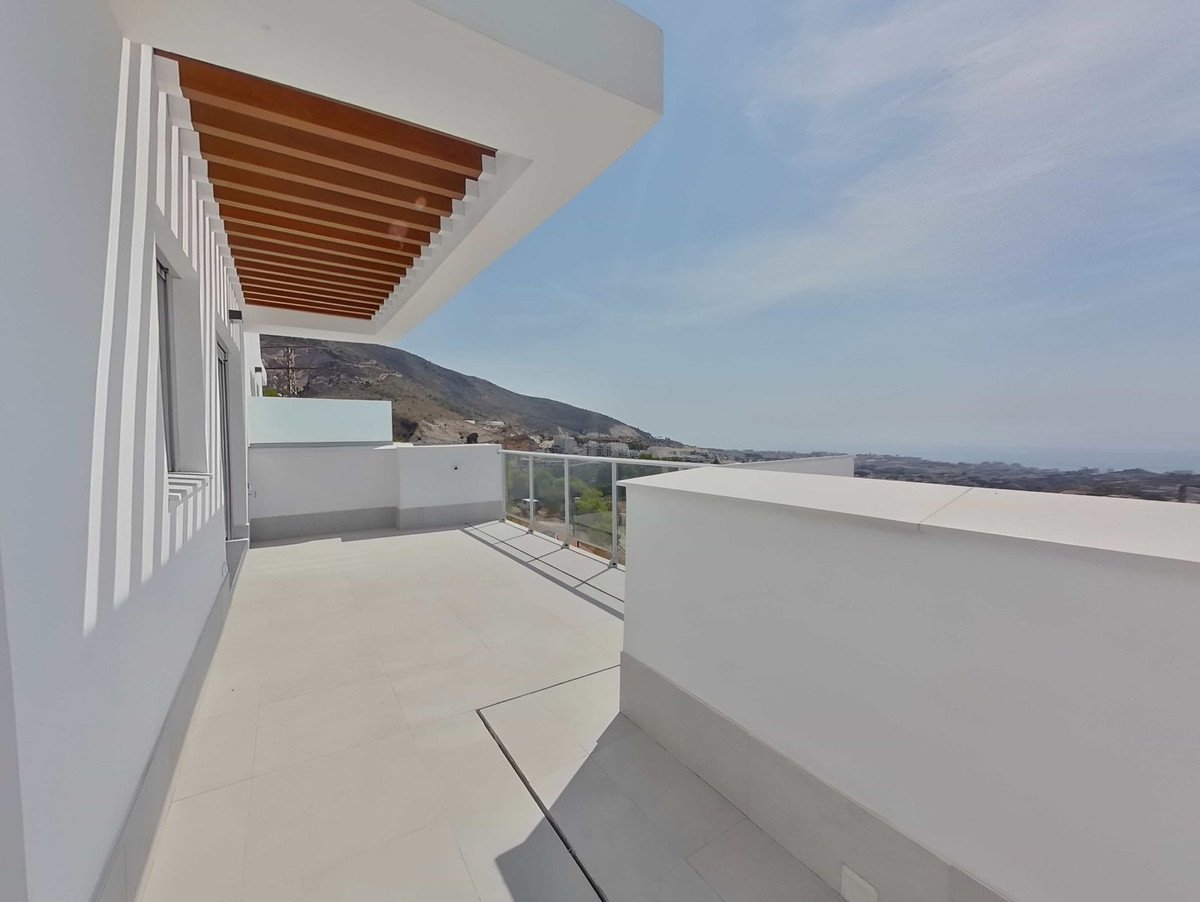 Spacious modern terrace with glass balustrade, wooden ceiling detail, and stunning sea and mountain views under a clear blue sky.