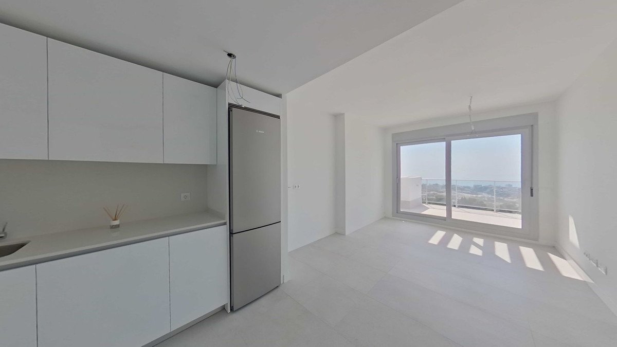 A modern kitchen area with white cabinets and a refrigerator, opening into a bright living space with a large window overlooking a balcony and distant landscape.