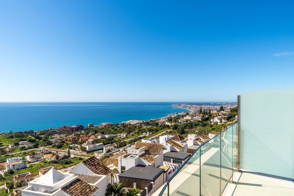 Spacious balcony with glass railing overlooking the sea, coast, and town below under a clear blue sky.
