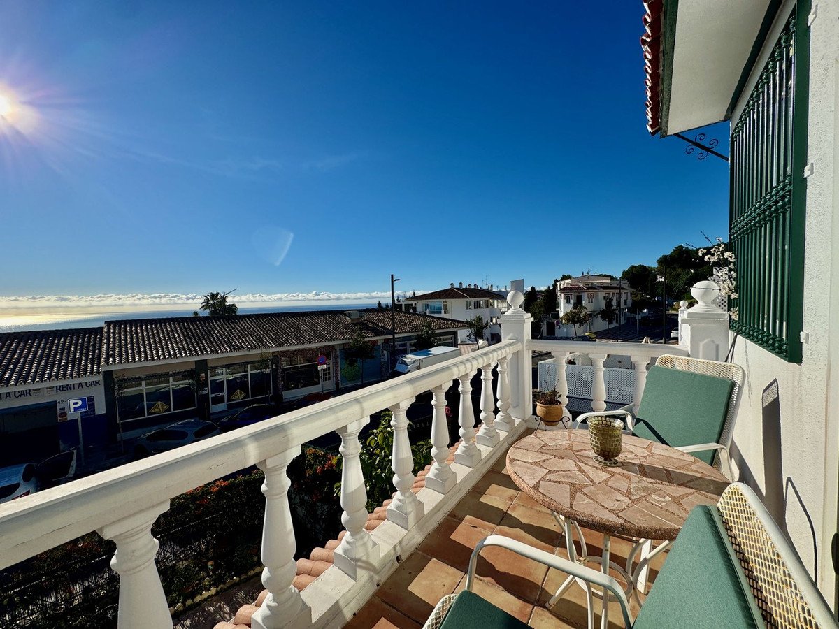 A balcony with a table and chairs offers a view of the sea and distant buildings under a bright sunny sky.