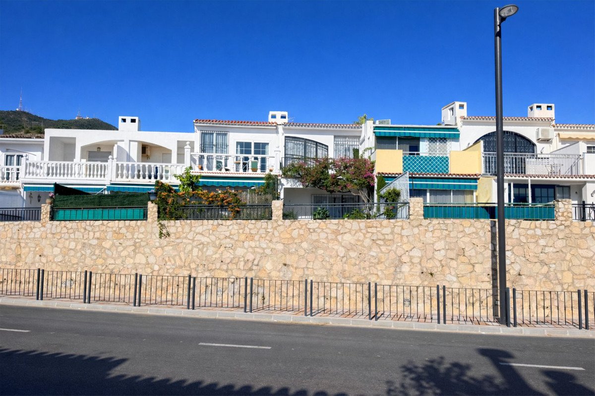 Row of terraced houses with white facades, green awnings, and balconies. A stone wall and a street are in the foreground.