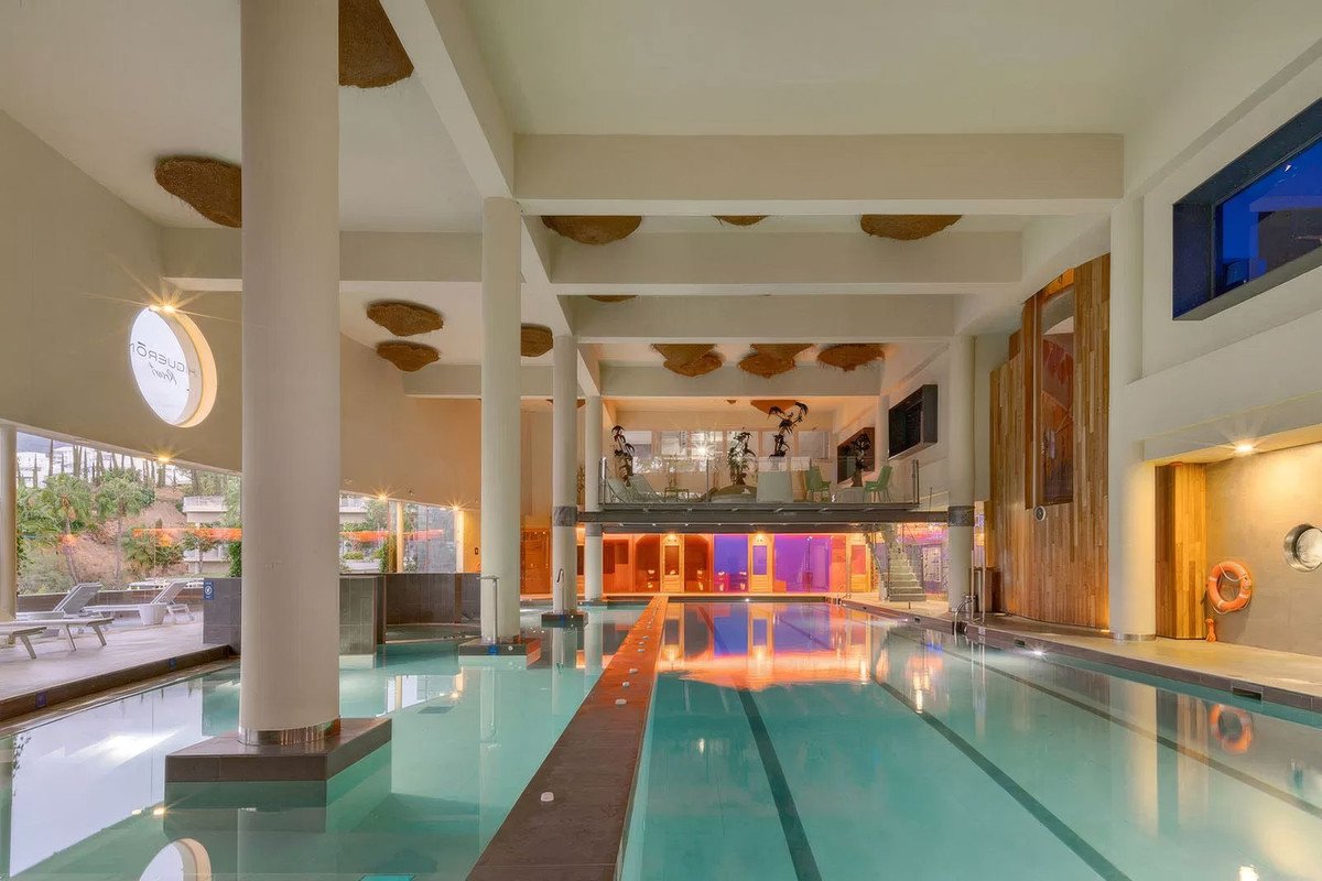 Modern indoor swimming pool with unique ceiling decor, featuring lounge chairs and a gym area on the upper level.