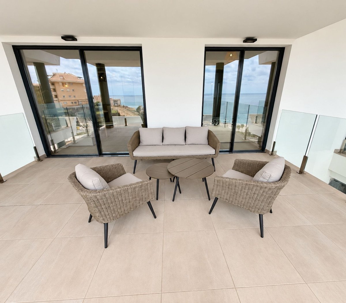 A modern terrace with woven seating furniture and coffee tables, offering a beautiful sea view through large glass doors.