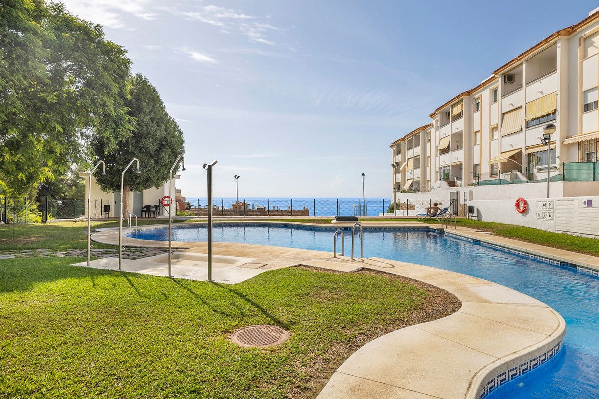 Outdoor swimming pool with showers, surrounded by green lawn and buildings, overlooking the blue sea under a clear sky.