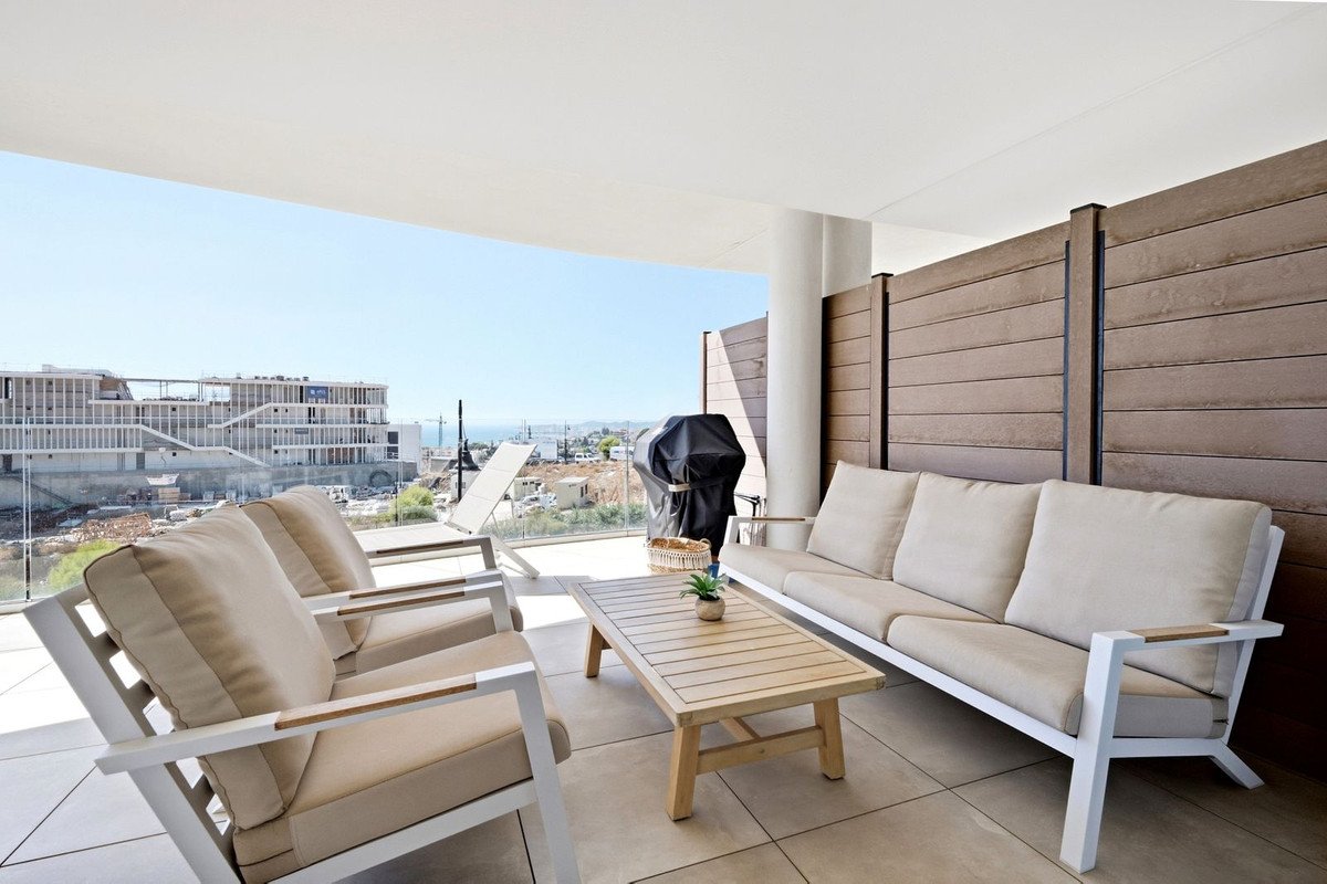 Modern patio with comfortable seating, coffee table, grill, and a view of the sea and buildings under construction.