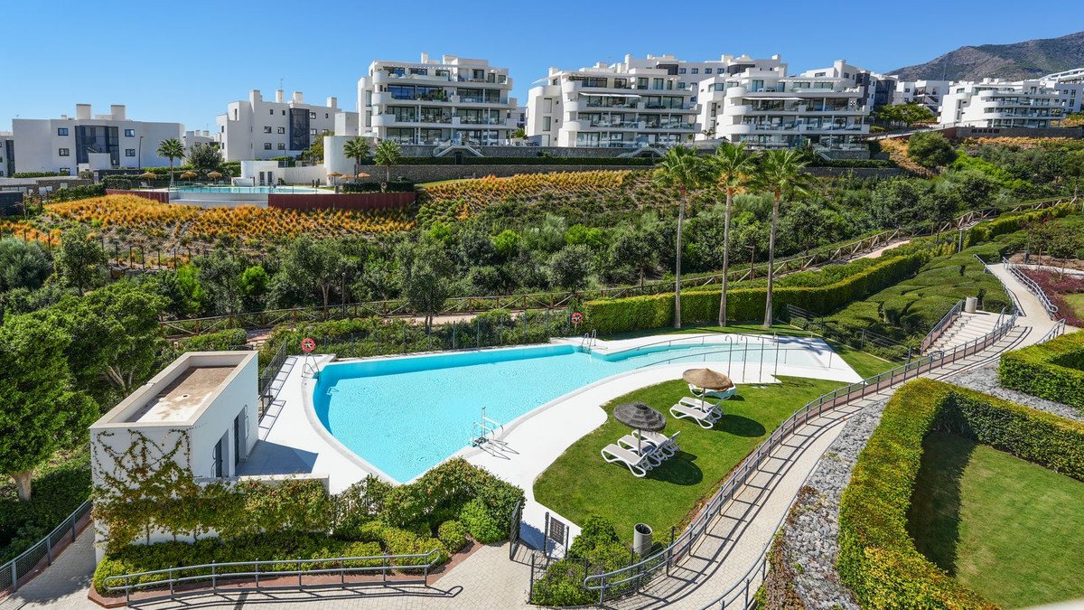A bright blue swimming pool with sun loungers and umbrellas, surrounded by green lawns and modern apartment buildings.