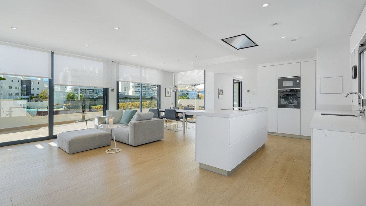 Modern open-plan living area with sofa, dining table, and a sleek white kitchen. Large windows offer a view of the pool.