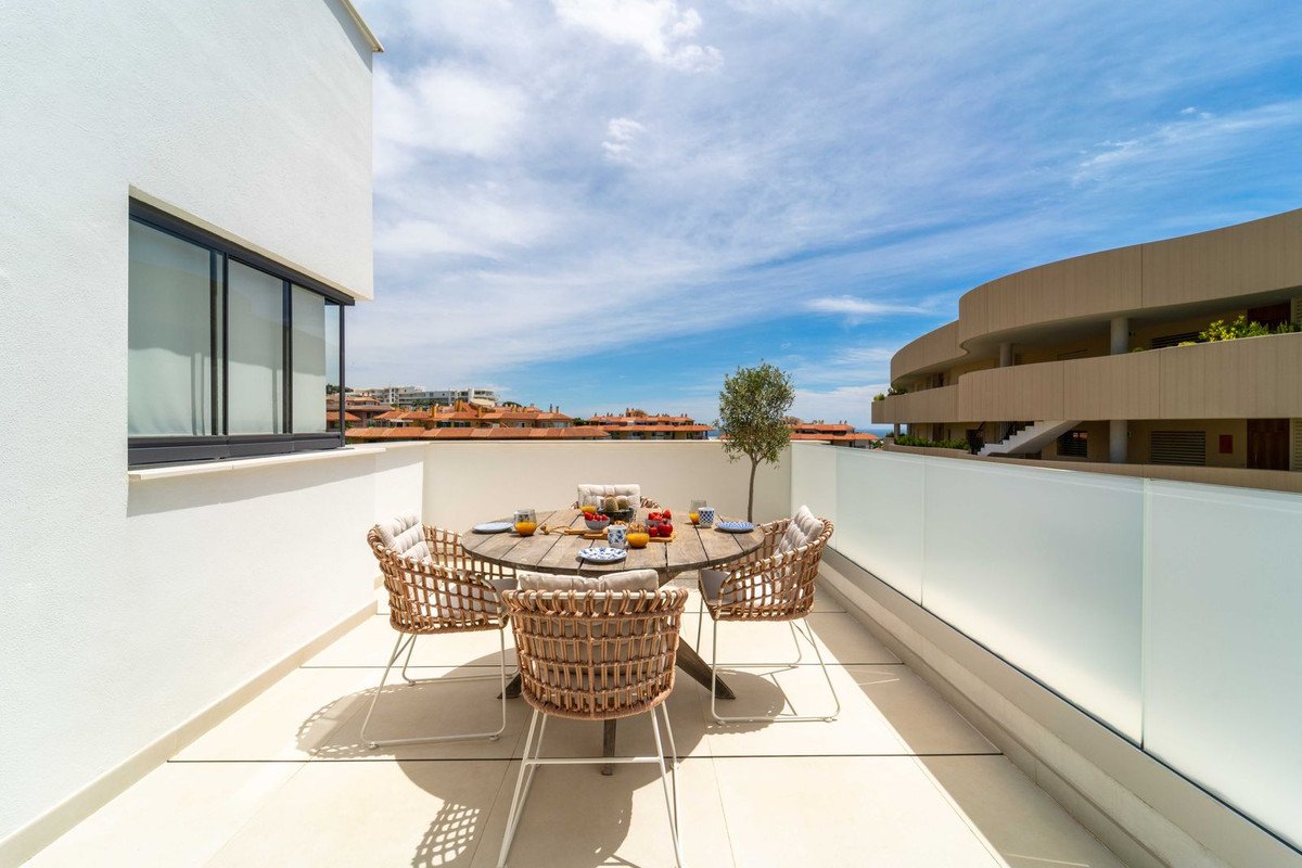 A furnished terrace with a dining set, overlooking rooftops and a cloudy blue sky, offering a distant sea view.