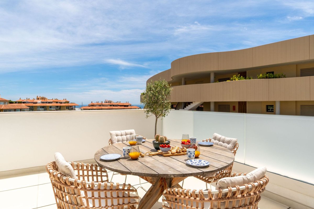 Rattan chairs around a wooden table set for breakfast on a sunny terrace with a sea view in the distance.