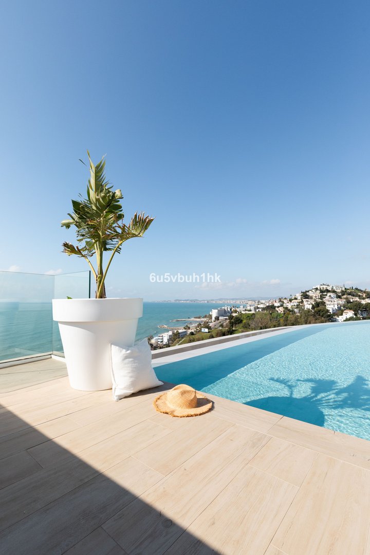 Infinity pool overlooking the sea and a coastal town, with a straw hat and potted plant on the deck.