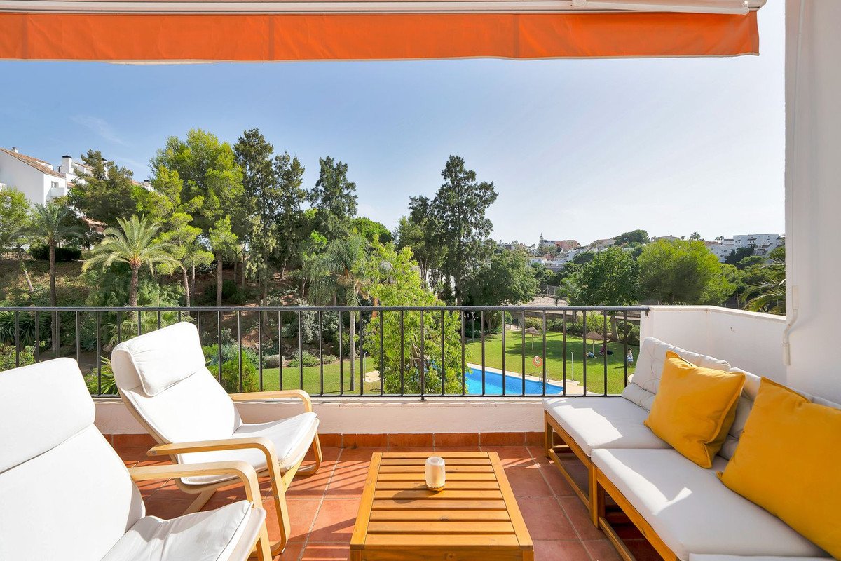 A sunny terrace with lounge seating, a coffee table, and views of a garden with a swimming pool and distant buildings.