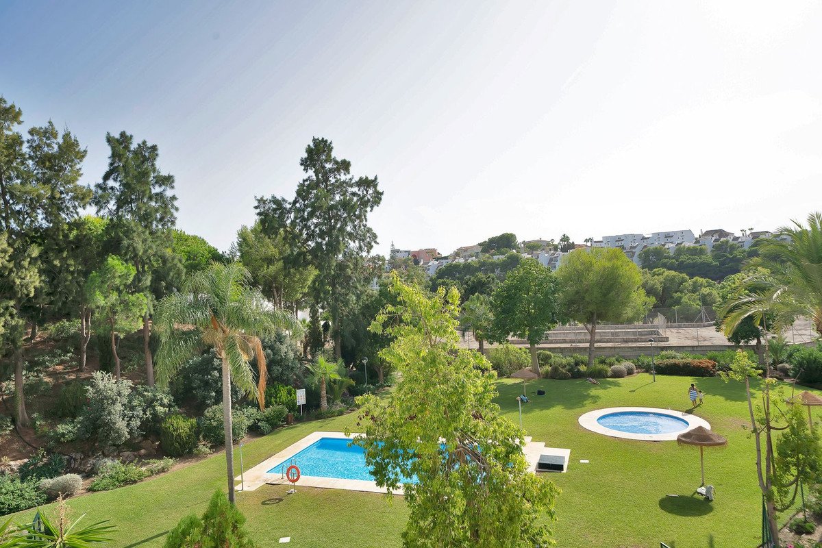 Outdoor swimming pools and manicured lawns surrounded by lush green trees and hillside buildings in a resort setting.