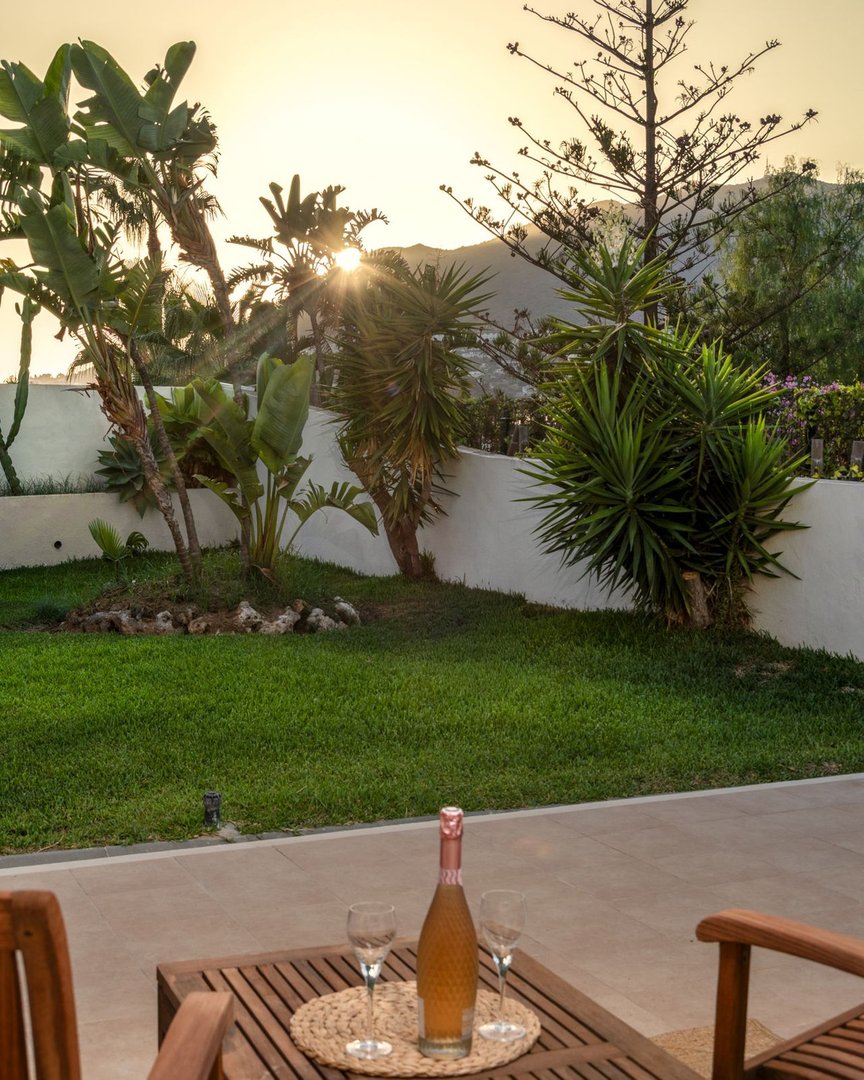 Bottle of wine and glasses on a patio table overlooking a garden at sunset with lush greenery.