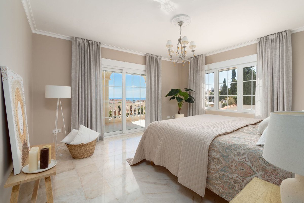 A bedroom with a large bed, balcony access with sea view and elegant decor. Sunlight streams in.