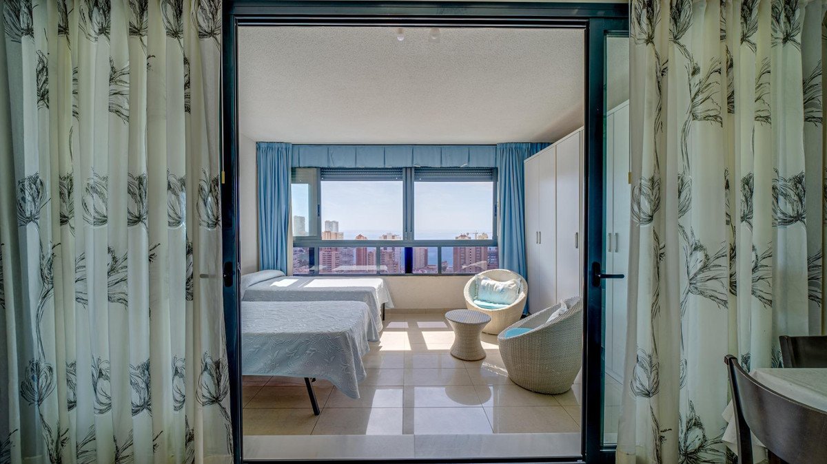 Spacious bedroom with two beds, balcony view of city and sea, wicker furniture, and floral curtains.