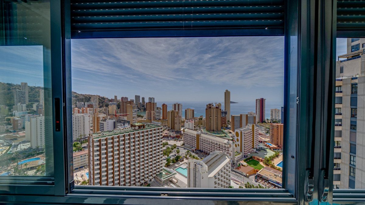 Balcony view of a cityscape with high-rise buildings and the sea, seen through a window with blinds.