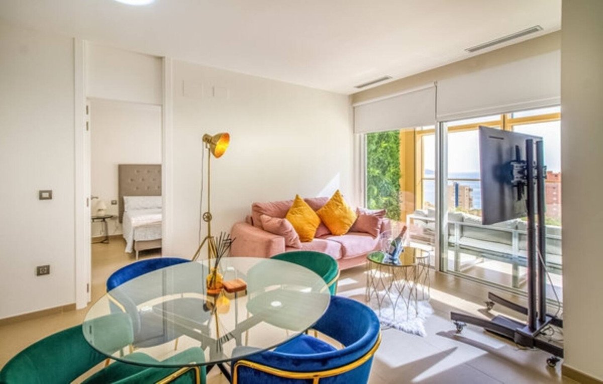 Modern living room with pink sofa, dining table, and balcony overlooking the sea and cityscape.