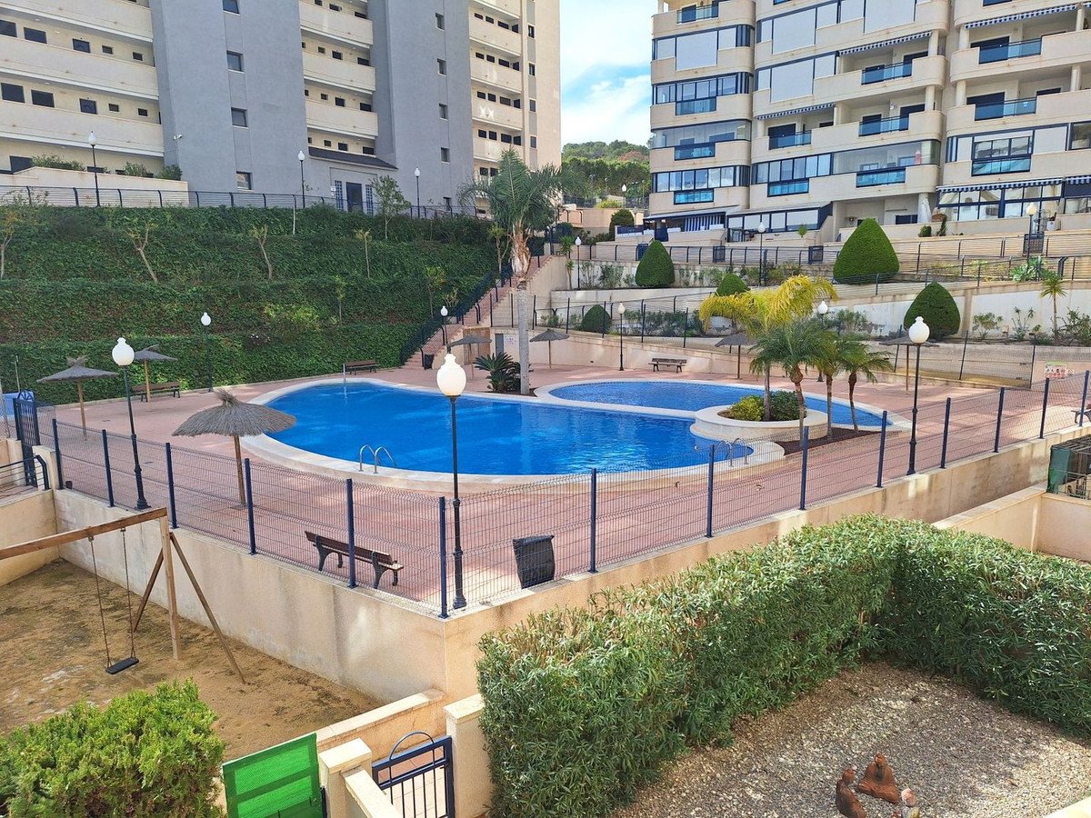 A bright blue swimming pool with a fence and umbrellas, adjacent to apartment buildings and a children's swing set.