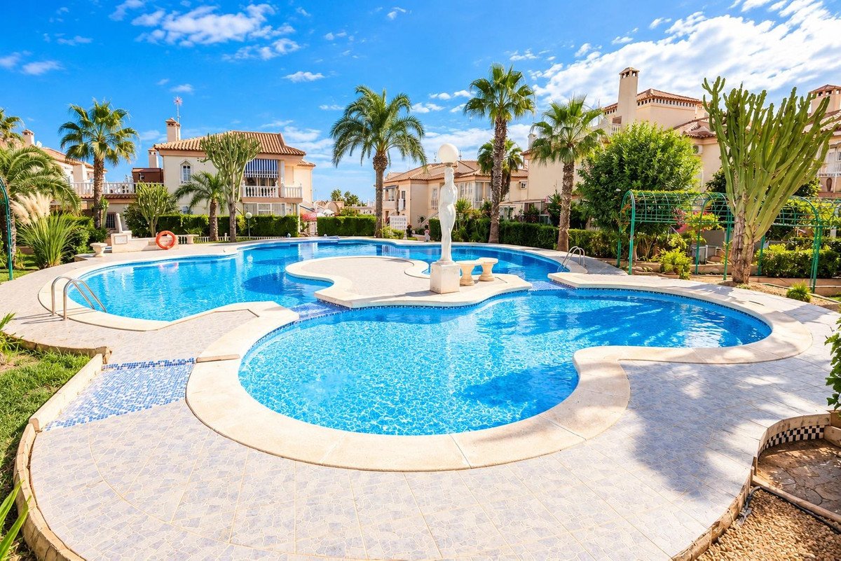 A large, multi-level swimming pool with palm trees and apartment buildings in the background on a sunny day.