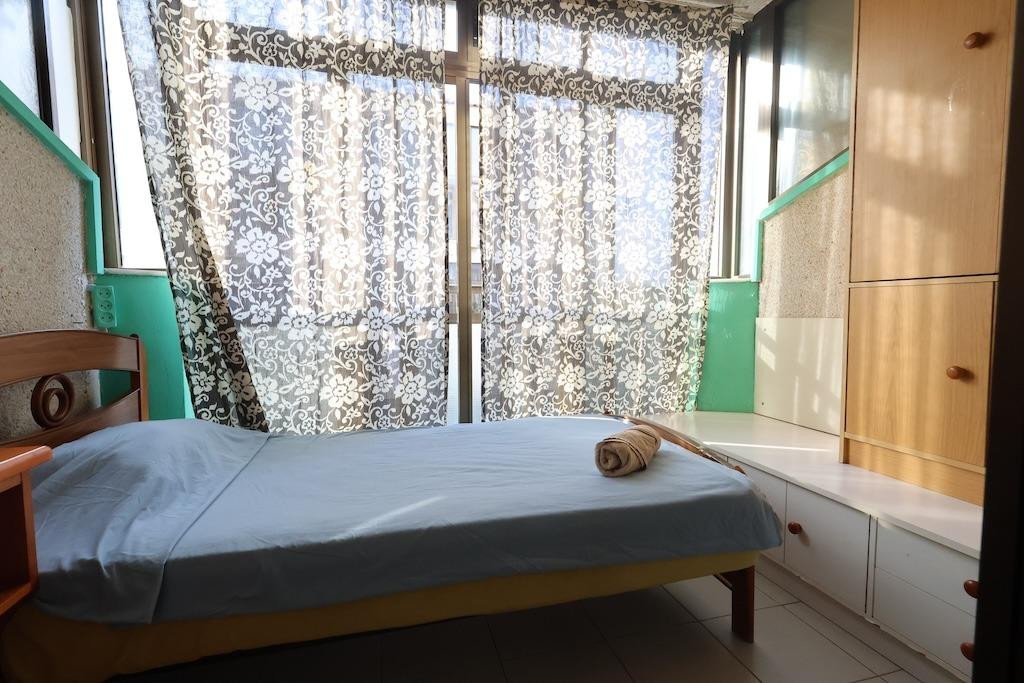 A neatly made bed with light blue sheets and a rolled towel, positioned in front of large patterned curtains and a wooden wardrobe.