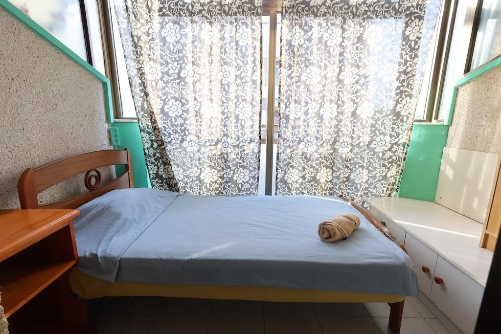 A bedroom with a double bed, blue sheets, and patterned curtains. A wooden bedside table is on the left.