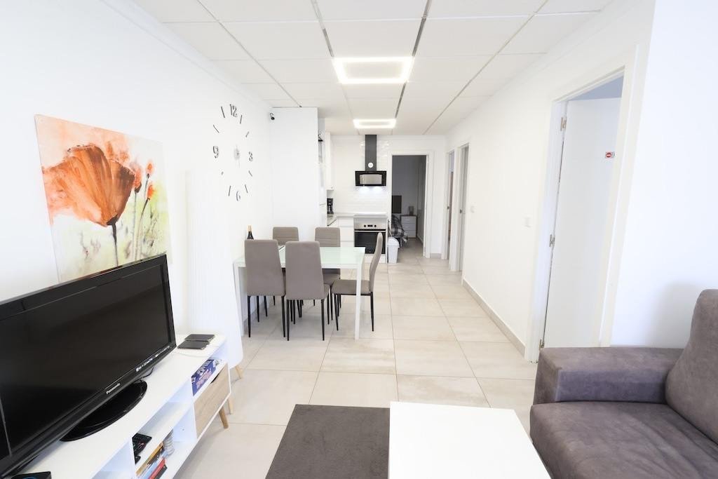 A spacious, modern living and dining area with a kitchen visible in the background. Features a TV, dining table, and sleek lighting.