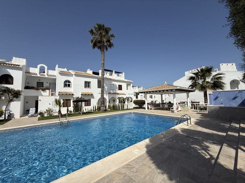 A long, blue swimming pool with a covered outdoor seating area and white buildings under a clear blue sky.