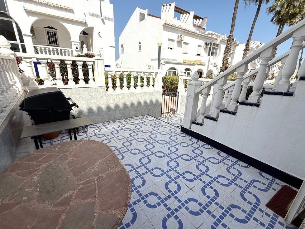 Outdoor terrace with patterned tiles, a grill, table, and stairs leading up to white buildings with palm trees overhead.