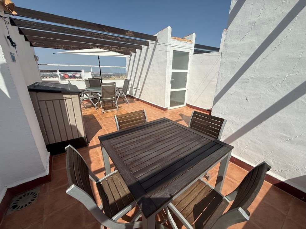 Terrace with wooden table and chairs, storage box, and a view of the horizon under a pergola with an umbrella.