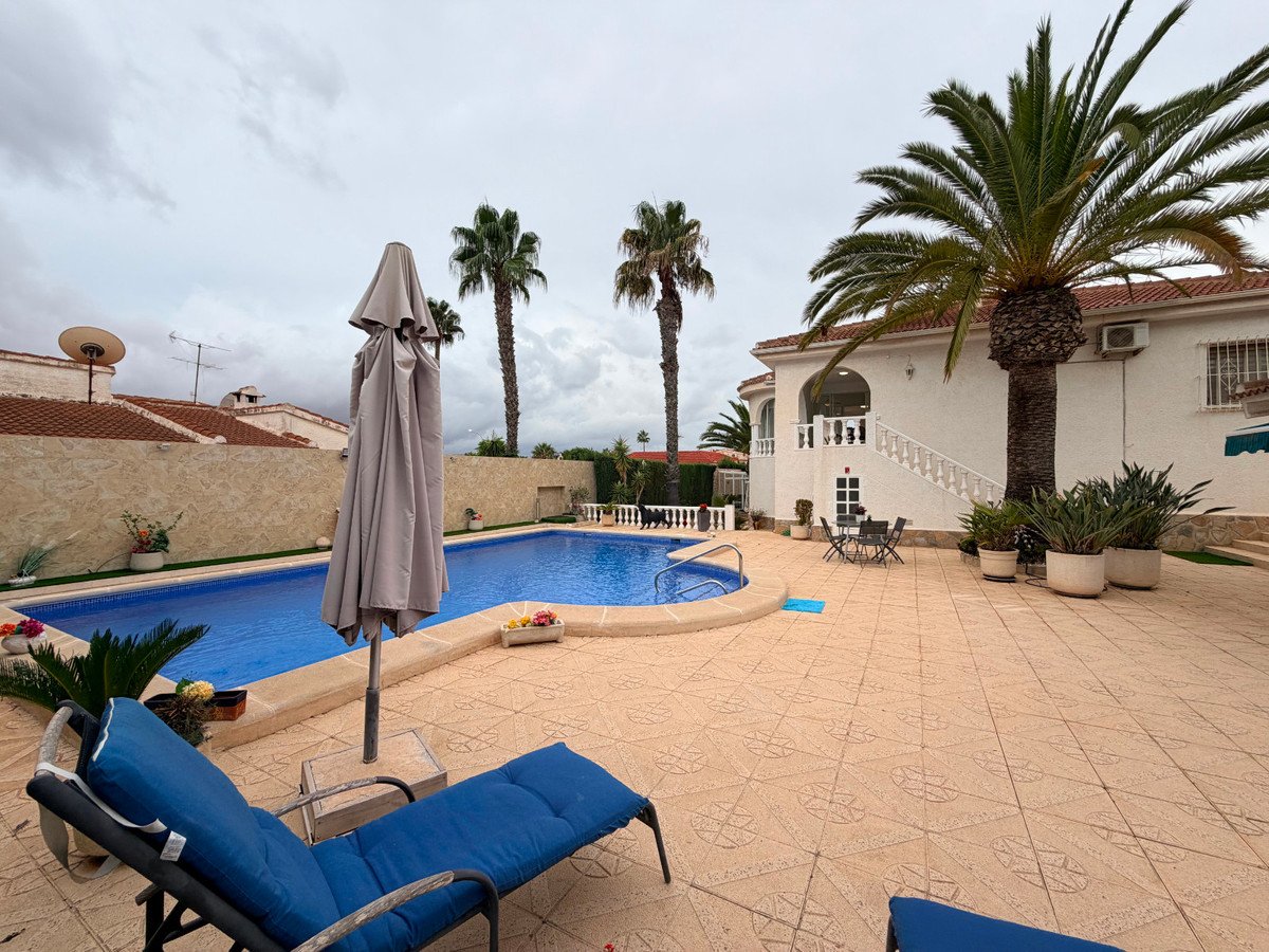 A patio with a swimming pool, lounge chairs, and palm trees in front of a white house.