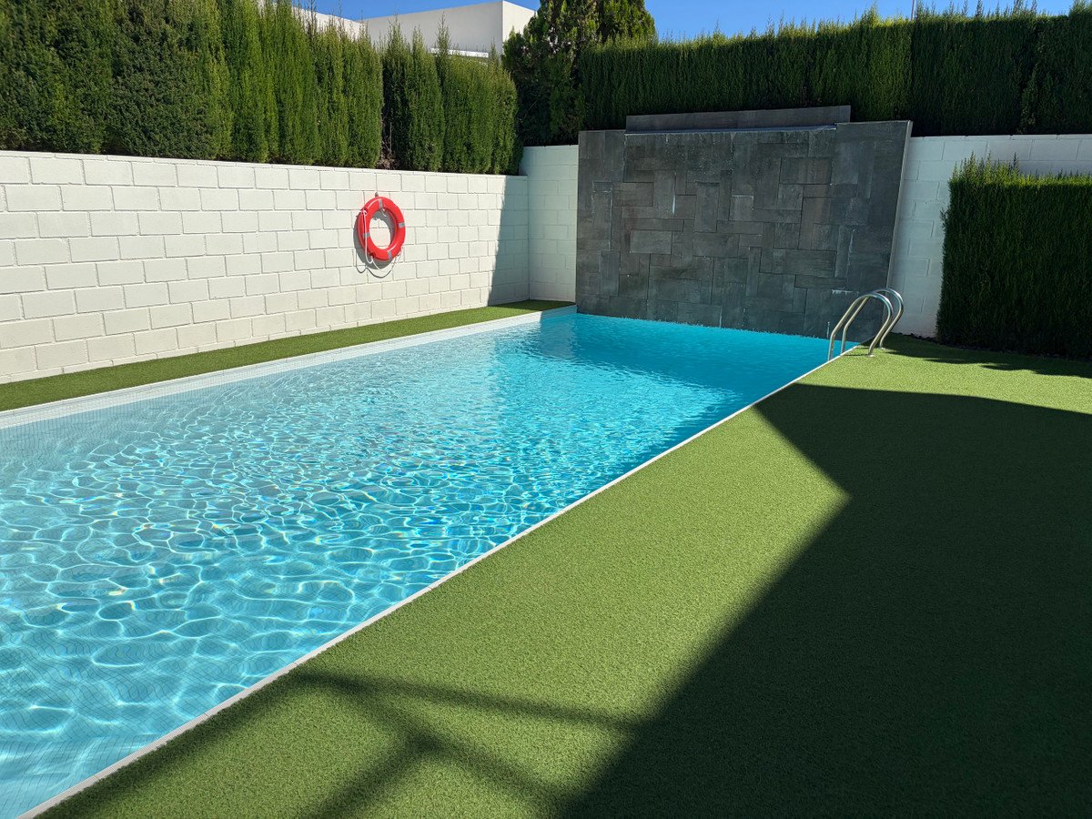 Rectangular swimming pool with a modern stone waterfall feature, surrounded by artificial grass and lush green hedges.