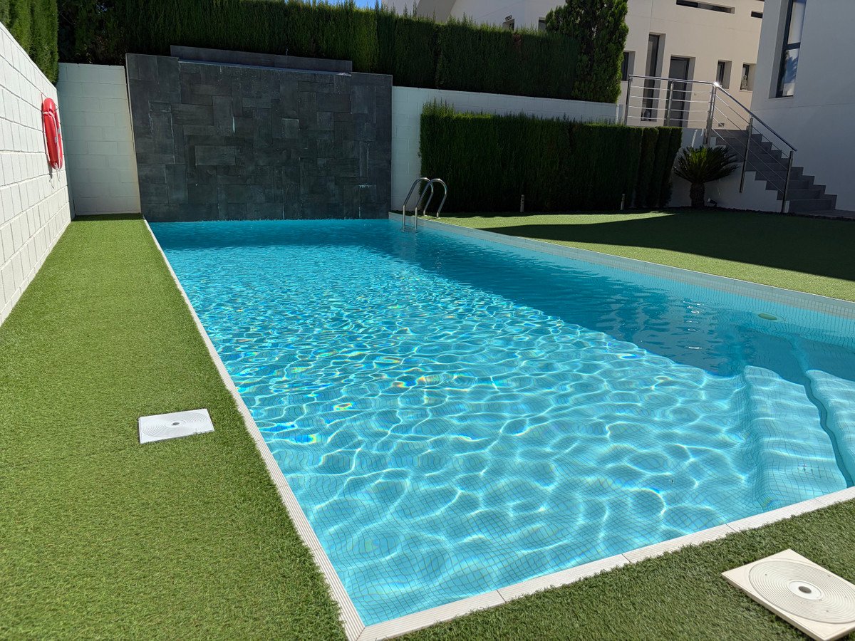 Modern swimming pool with a dark stone waterfall feature, surrounded by artificial turf and overlooking a building and manicured hedges.