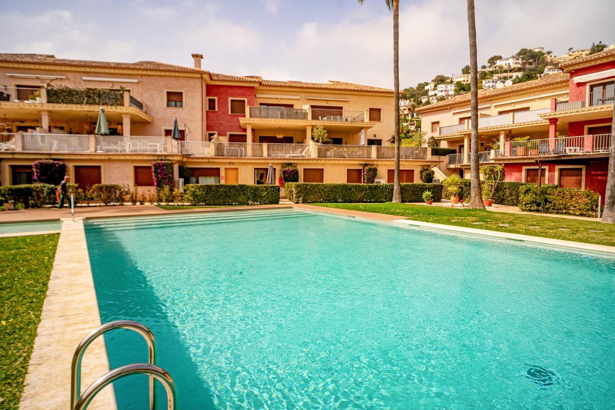 A large, bright blue swimming pool in front of a residential building with balconies and manicured hedges under a clear sky.