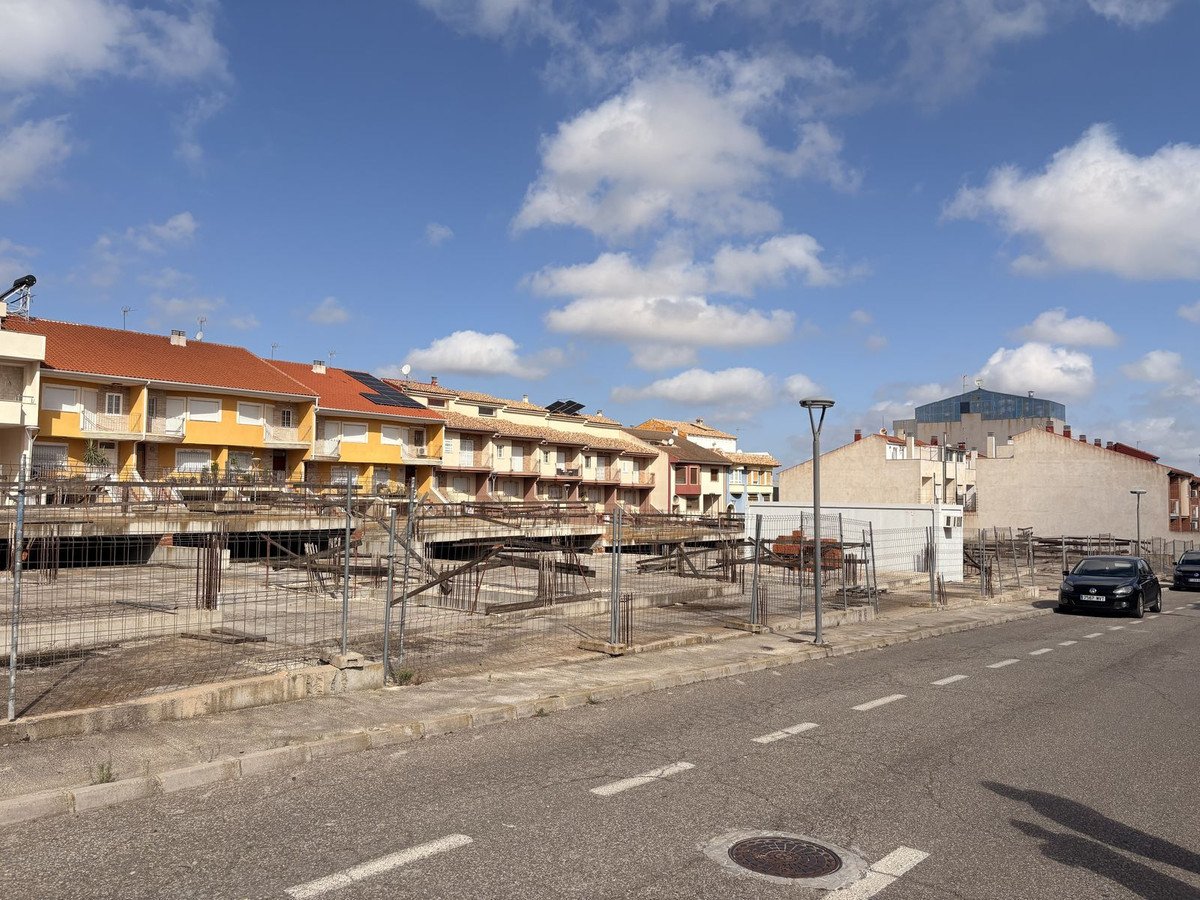 Construction site in front of residential buildings under a blue sky with clouds. Site is fenced off with visible rebar and concrete structures.