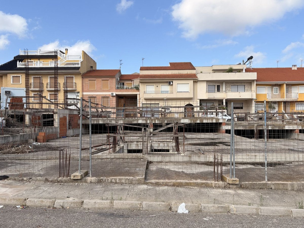 Construction site with rebar and concrete foundations in the foreground, residential buildings in the background under a blue sky with clouds.