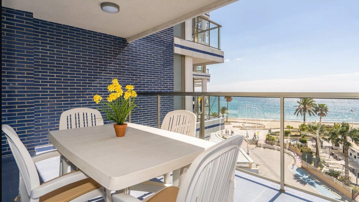 Balcony with white table and chairs, yellow flowers, overlooking beach and ocean with palm trees.