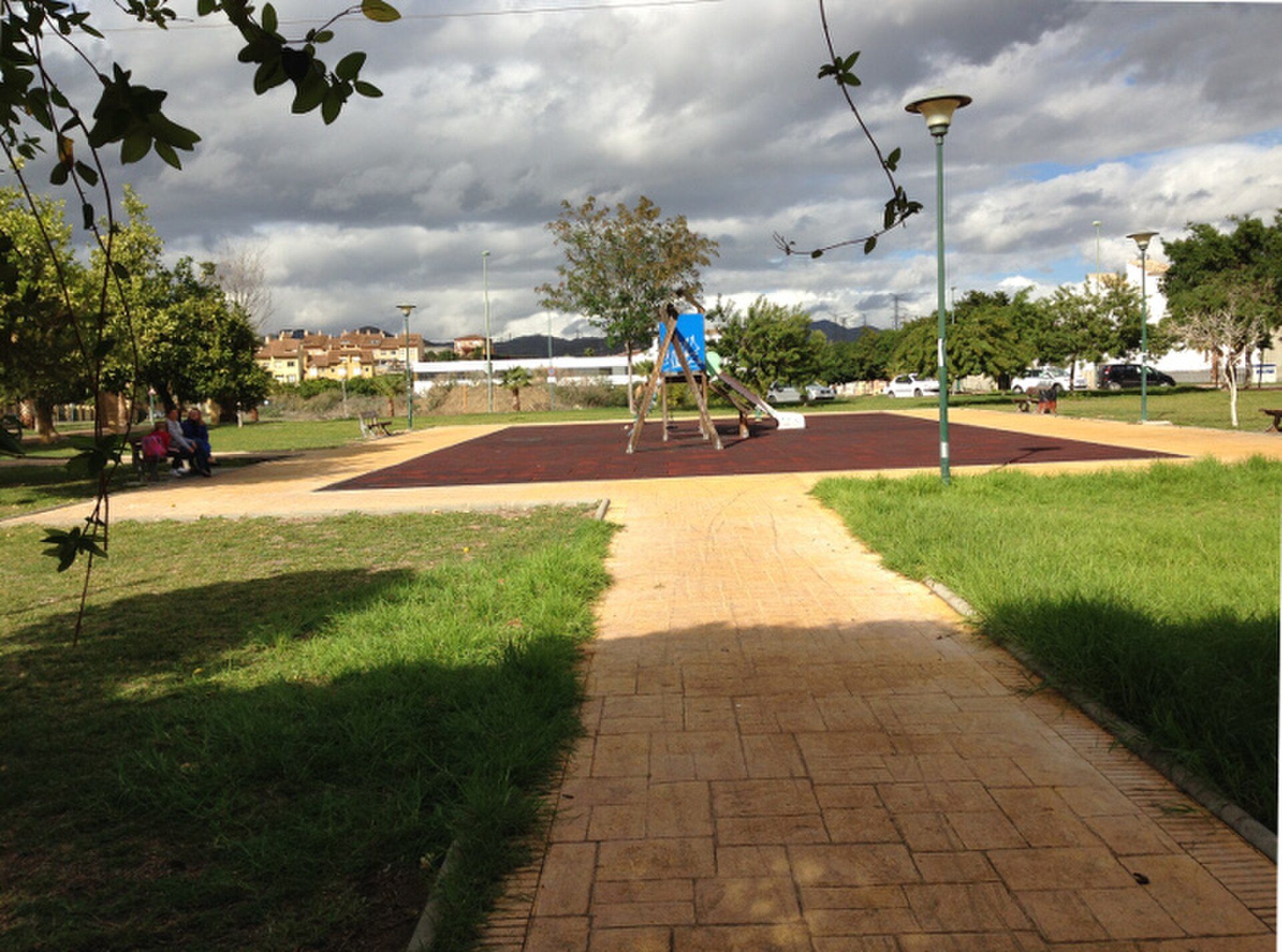 A park with a playground, grassy areas, and a paved path under a partly cloudy sky.