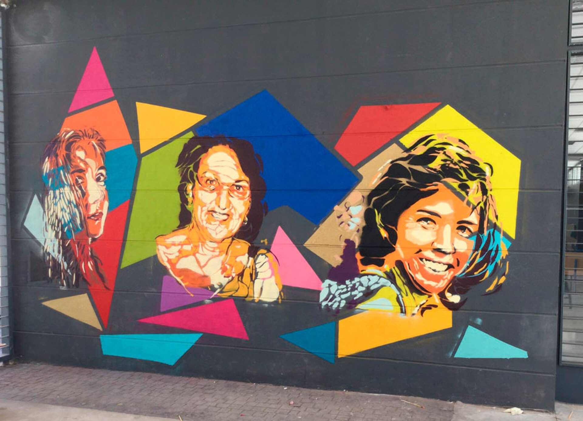 Colorful mural with abstract shapes and three faces on a dark wall.