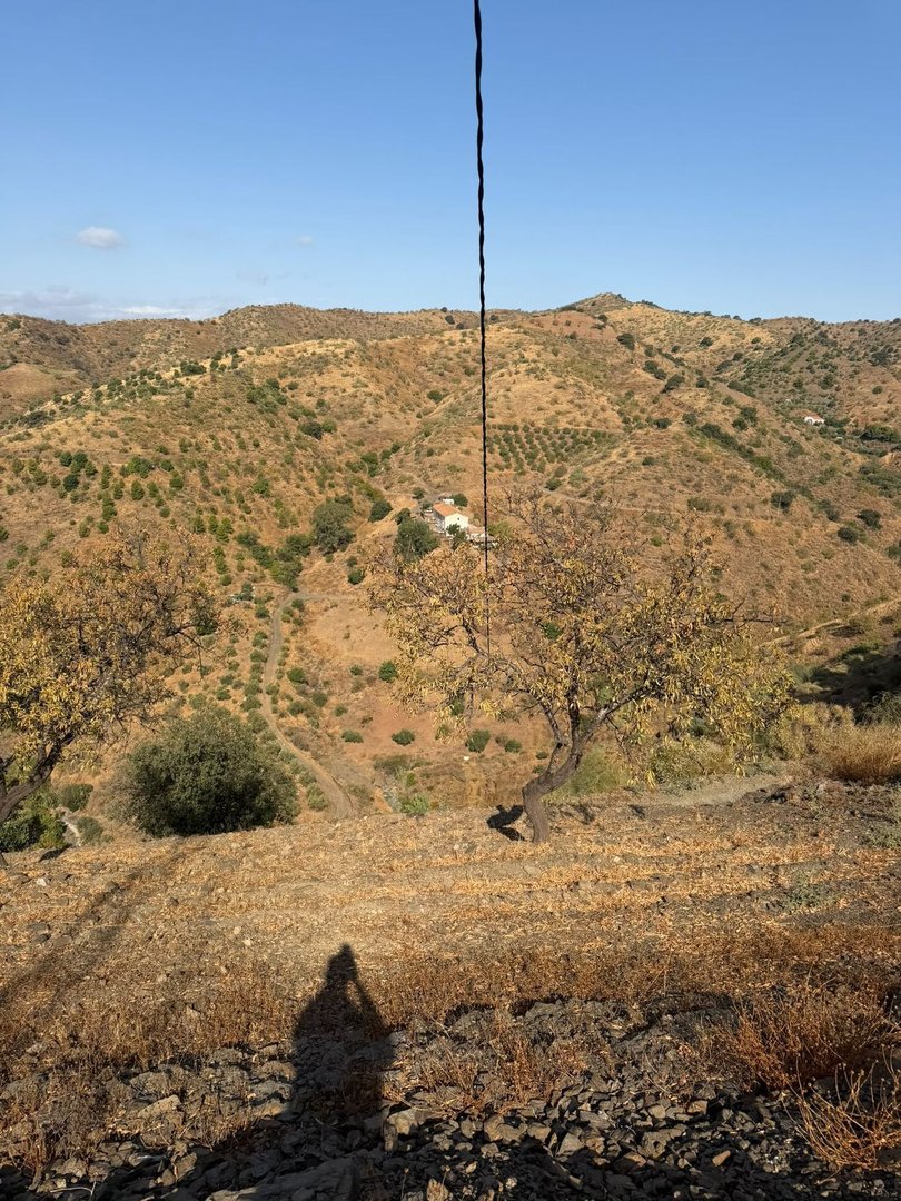 A rural landscape with rolling hills, scattered trees, and a distant house under a clear blue sky.