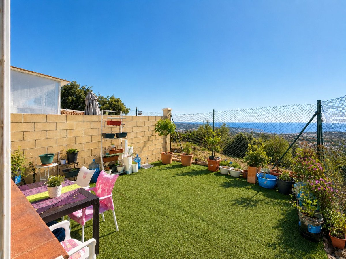 A garden terrace with artificial grass, potted plants, a dining table, and a view of the sea and hills under a clear blue sky.