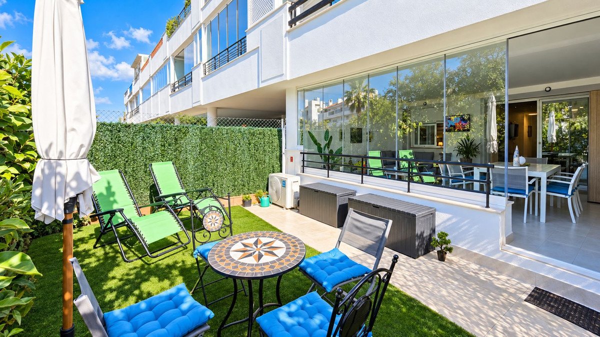 Sunny garden terrace with green lounge chairs, mosaic bistro set, and glass-enclosed dining area visible through large windows.