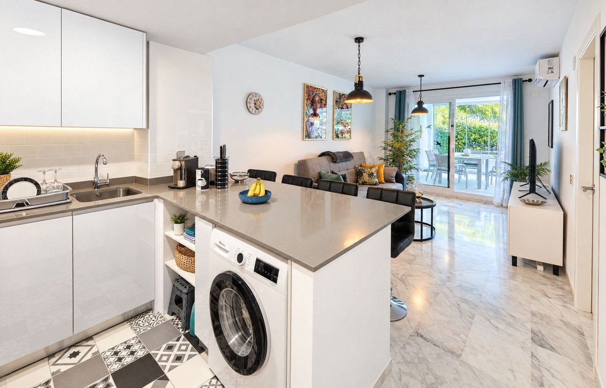 Modern kitchen island with washing machine, open living area, and sliding doors leading to a furnished terrace with garden views.