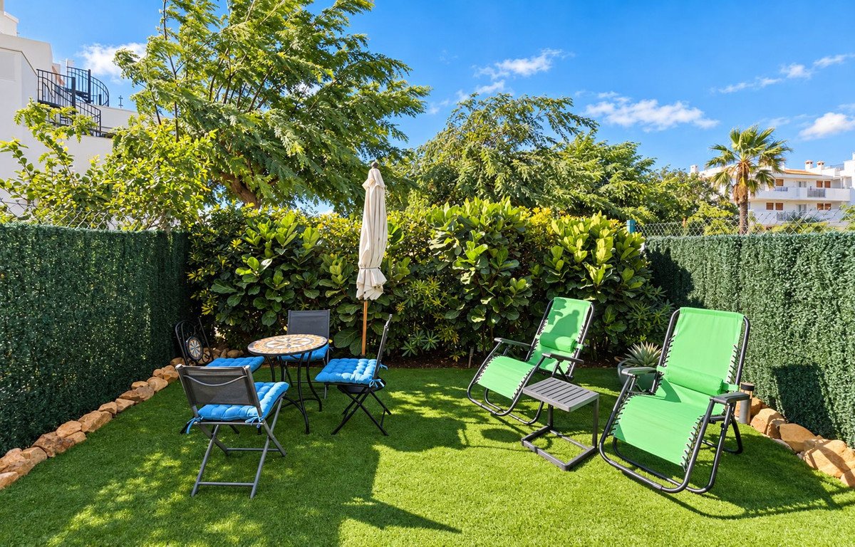 Artificial lawn backyard with two green lounge chairs, a small bistro set, and a closed umbrella under a blue sky.