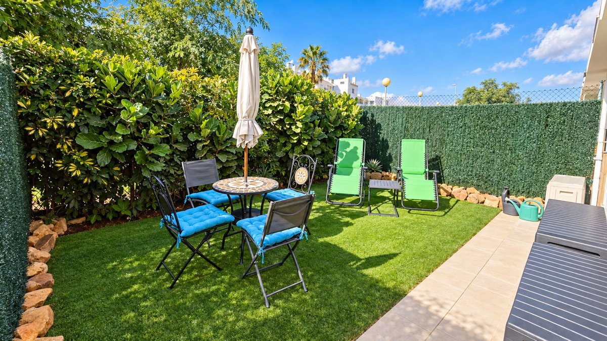 A sunny garden patio with green artificial turf, seating for four, lounge chairs, and a small table with an umbrella.