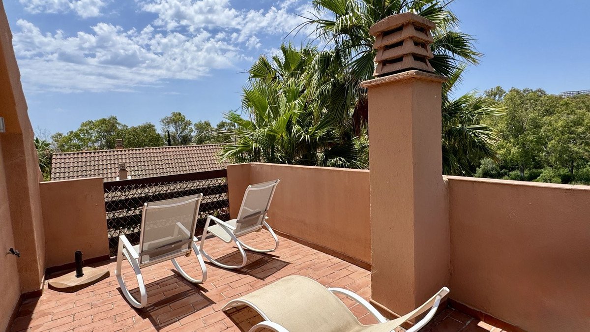 Terrace with chairs and lounge bed, overlooking palm trees and greenery under a blue sky with clouds.