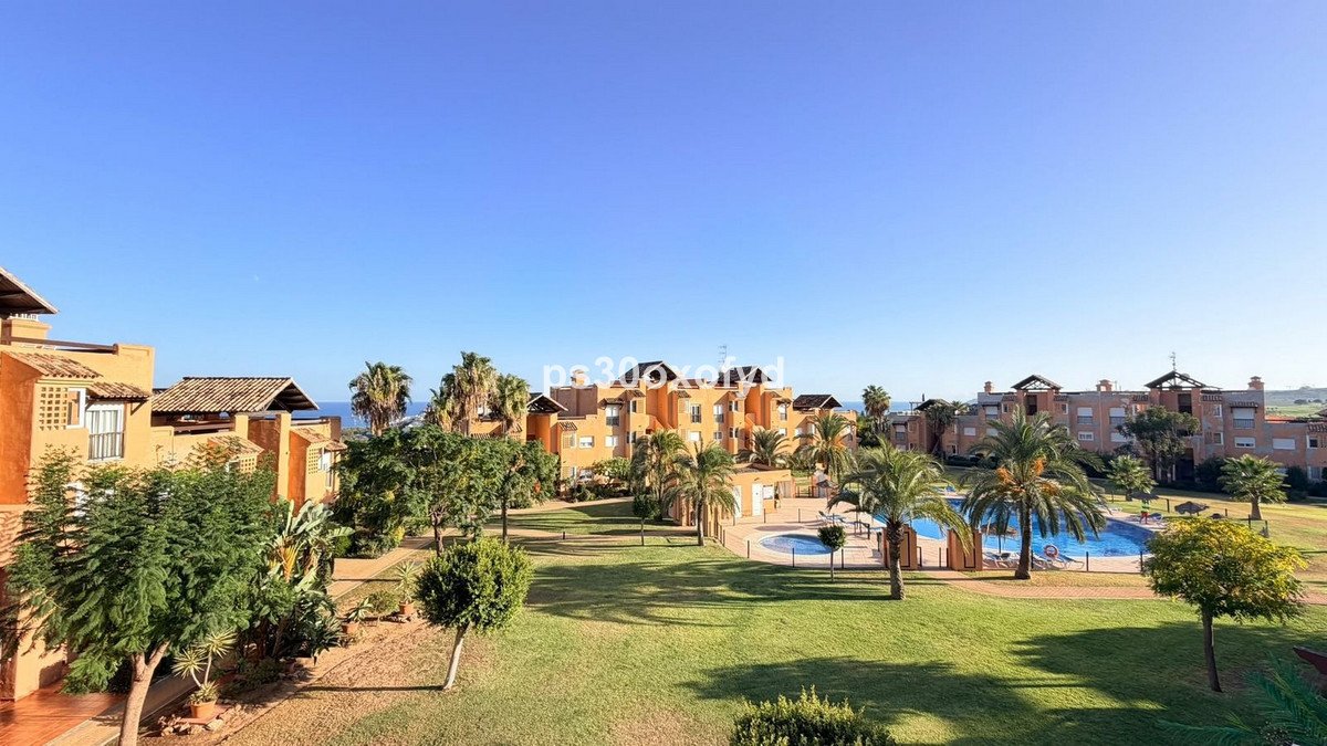 An exterior view of an apartment complex with a swimming pool, lush green gardens, palm trees, and a distant view of the sea under a clear blue sky.
