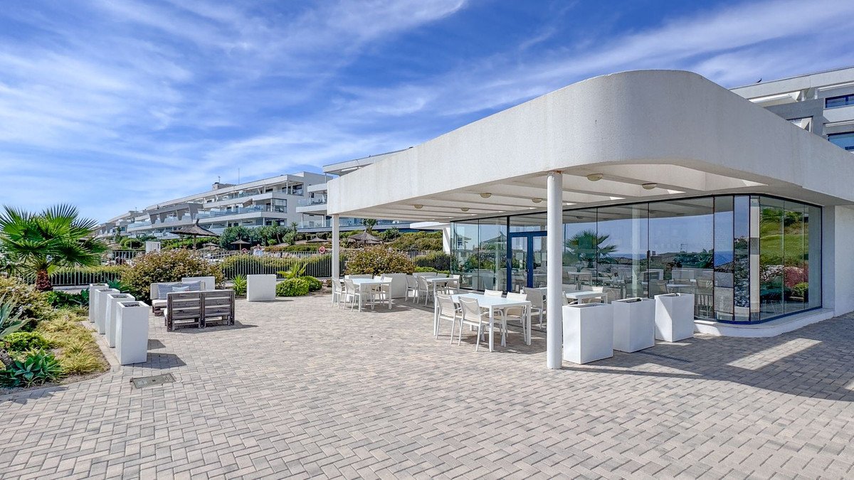 Outdoor dining area with white tables and chairs overlooking a modern building and the sea under a blue sky.