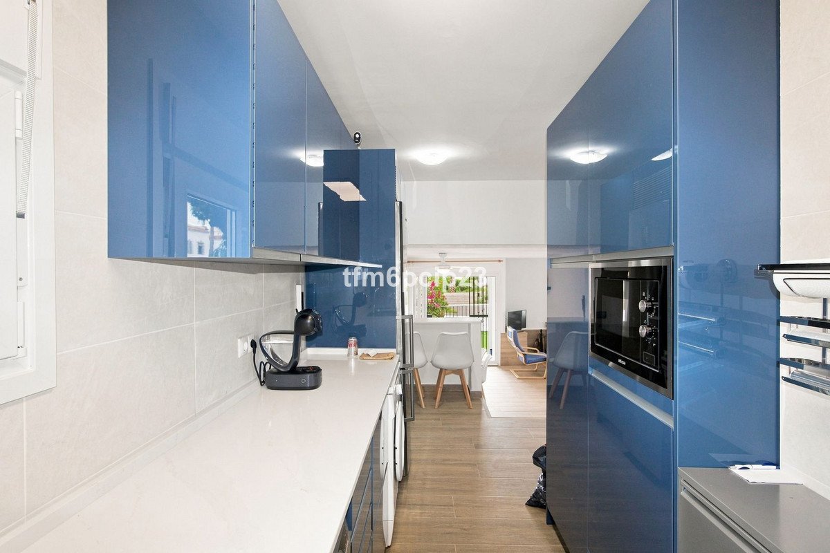A modern kitchen with high-gloss blue cabinets and a white countertop, opening to a living area with furniture and a glimpse of greenery outside.