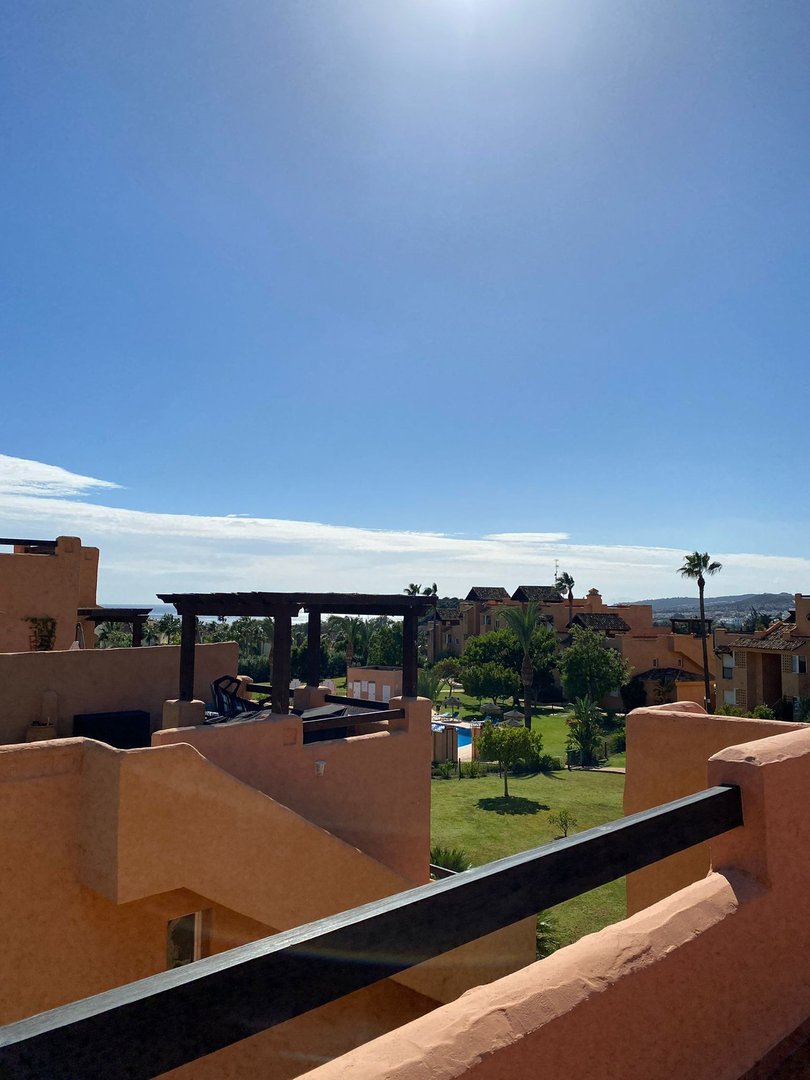 Terrace view overlooking a resort with a pool, green lawn, and distant buildings under a clear blue sky.