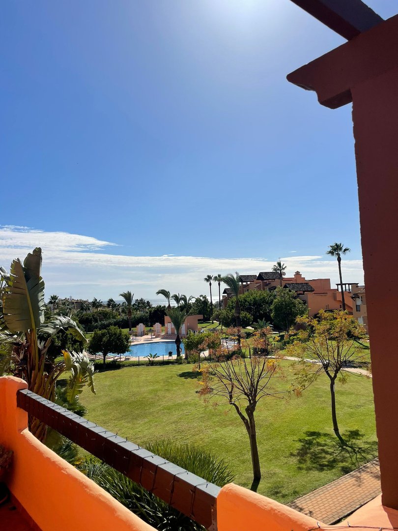 View from a balcony overlooking a lush green garden with a swimming pool and palm trees under a clear blue sky.